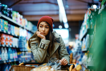 Young woman feels worried about increase in food prices while buying in supermarket.