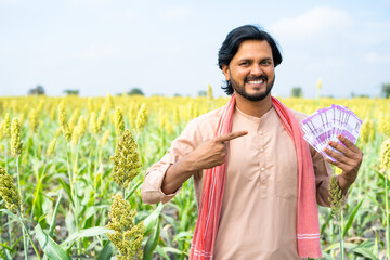 Portrait shot of Happy Indian farmer showing money by pointing finger while looking camera at farmland with copy space - concept of earning, agriculture profit and successful investment © WESTOCK