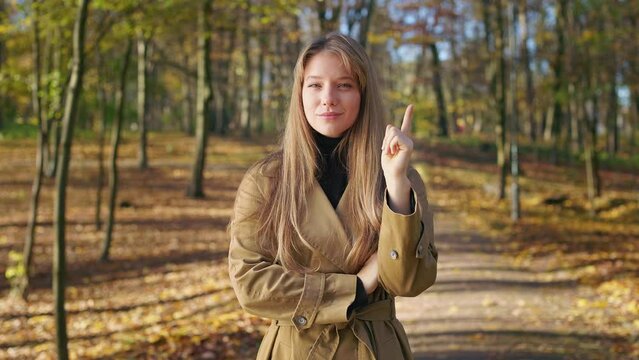 Front view of elegant, young woman walking in park, standing. Beautiful lady looking at camera, raising hand, finger, squint, remembering, smiling. Concept of modern lifestyle.
