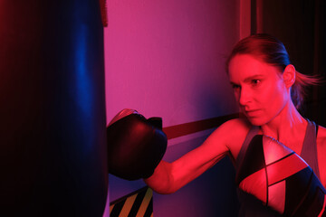 Woman punching a bag with boxing gloves illuminated by neon pink light.