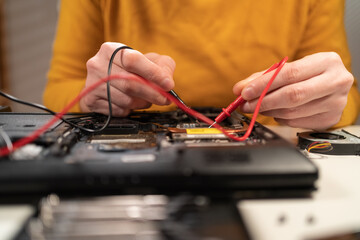 Close-up of man hands testing and examining laptop motherboard with multimeter at desk. Maintenance, repairing and checking computer hardware