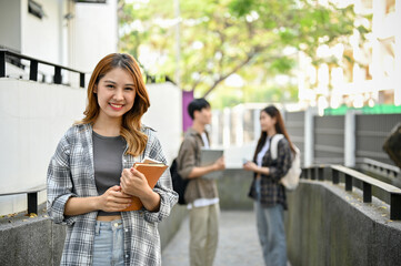 Attractive Asian female college student with her school book stands in front of the campus building.