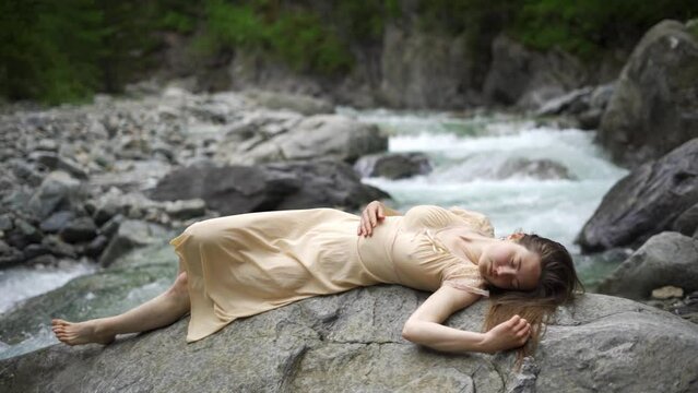 Young attractive woman in a wet water color dress lies on the stones. A tender, fragile, slender, river nymph sleeping beauty, resting near a tropical waterfall.