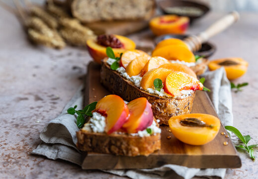 Open sandwiches with tartine bread and cream cheese, nectarine and apricot drizzled with honey on wooden cutting board. Concrete background.