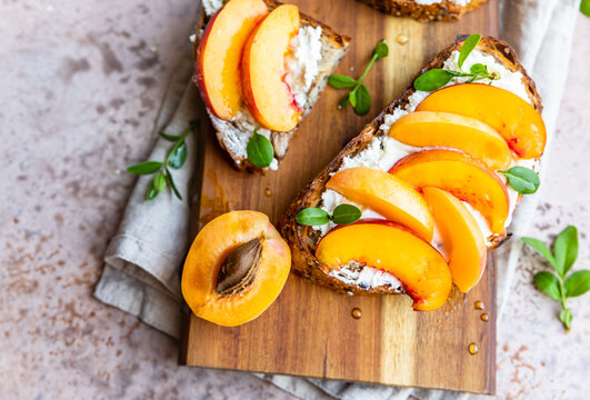 Open sandwiches with tartine bread and cream cheese, nectarine and apricot drizzled with honey on wooden cutting board. Concrete background.