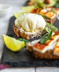 Variety of sandwiches with tartine bread with poached egg, mashed avocado and grilled cheese with salad leaves and lime on slate board. Healthy breakfast or brunch. Diet food concept.