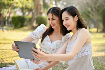 Two beautiful young Asian women are using tablet, taking selfies while picnic in the park.