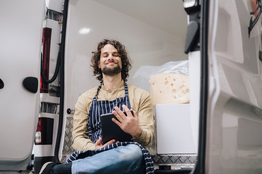 Smiling Young Entrepreneur Sitting With Eyes Closed Holding Tablet PC In Van