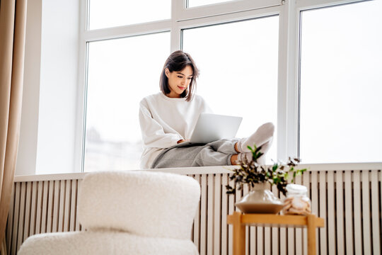 Smiling woman using laptop sitting on window sill at home
