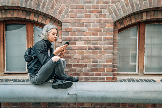 Woman sits cross-legged near brick building and looks at phone screen