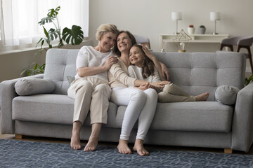 Cheerful loving child girl, mom and grandma sitting together on couch, enjoying family leisure, relaxation, meeting, hugging with affection, tenderness, care, smiling, chatting, laughing