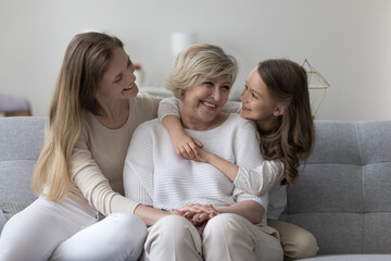 Sweet kid girl and mom visiting happy grandma, sitting on home sofa together, chatting, laughing, having fun, hugging with affection, enjoying family holiday, affection, relationship - obrazy, fototapety, plakaty