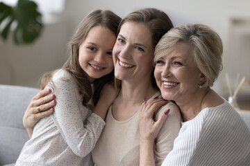 Three happy girls and women of different female family generations posing together for shooting, looking away with toothy smiles, laughing, resting on home sofa, enjoying leisure, having fun