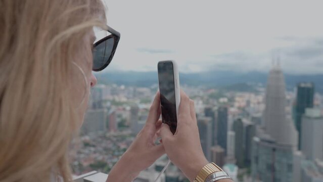Tourist woman on roof of skyscraper shoots a city landscape on a mobile phone. Rear view of lonely girl in twiligh on vacation. Kuala Lumpur capital of Malaysia.