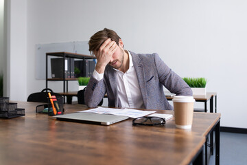 tired upset businessman sits in front of the desktop in the office covering his face with his palm