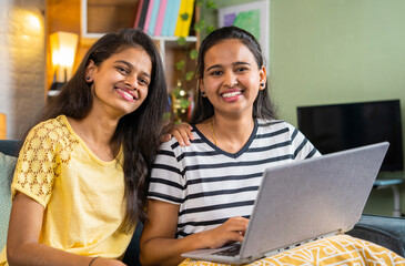 Happy smiling siblings girl using laptop by looking at camera at home - concept of technology, learning and bonding