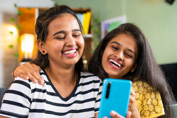 happy sibling girls laughing while using mobile phone on sofa at home - concept of entertainment, humour and positive emotion.