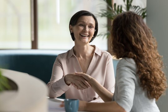Positive senior business woman giving handshake to younger colleague at work table, smiling, laughing, thanking for successful cooperation. Happy professional shaking hands in office