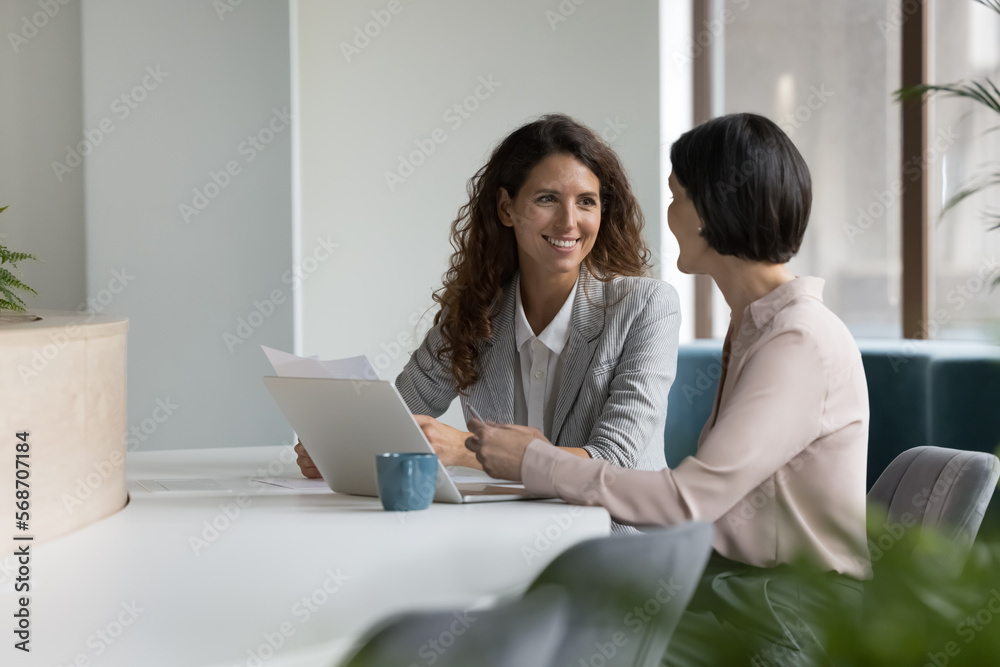 Wall mural two positive confident diverse business colleagues women talking at workplace, discussing project do