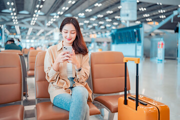 Happy asian tourist woman using mobile smartphone with suitcase traveling between waits for flight...