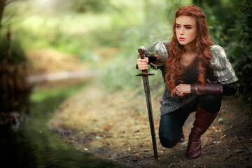 Beautiful red haired girl in metal medieval armor dress with sword standing in warlike pose and...