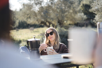 Blonde woman with sunglasses drinking a cup of coffee outside her camper van. Nature vacation...