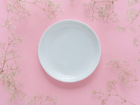 Ein wei&szlig;er leerer Teller umrahmt von Trockenblumen auf einem rosa Hintergrund. Kreis, Textfreiraum.