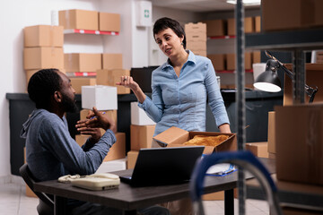Diverse employees discussing transportation logistics while preparing packages, putting customer order in cardboard box during job shift. Team working in delivery department in warehouse