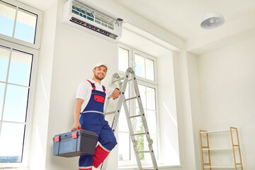 Male technician worker in uniform repairing air conditioner indoors. Air conditioning master standing on ladder with toolbox while installing or repairing conditioner in apartment