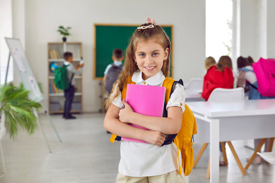 Portrait of happy little elementary school girl posing in classroom after lessons. Cute Caucasian girl 6-7 years old with backpack and notebook smiling looking at camera. Education and school concept. - Powered by Adobe