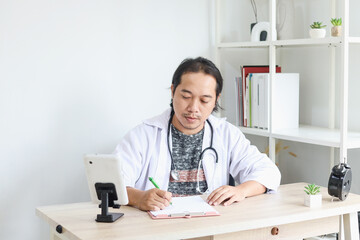 Obraz na płótnie Canvas Asian doctor sitting and writing on paper at clinic during online consultation. 