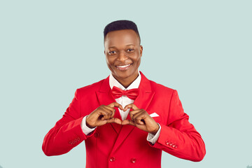 Handsome adult Afro American man in red suit makes hand heart shape. Benevolent human gentleman philanthropist businessman benefactor charity fund founder donates money wishes healthy life and harmony - obrazy, fototapety, plakaty