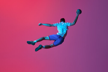 Fototapeta na wymiar Back view. In a jump. Young man, professional handball player training, playing isolated over gradient pink background in neon light. Concept of sport, action, motion, championship, sportive lifestyle