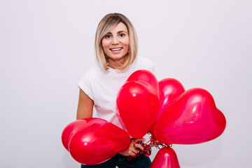 Excited Happy Girl with red hearts shape balloons in white interior near the door at home. Easy-going woman with blond hair expressing positive emotions in valentines day.