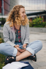 University student using smartphone app sitting outdoors watching webinar distance course or listening podcast. Millennial female texting and browsing internet