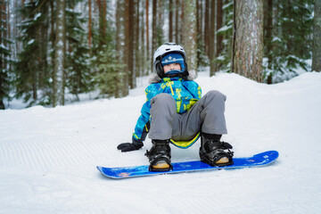 Fototapeta na wymiar little boy sitting on snow putting his feet in snowboard bindings adjusting straps.