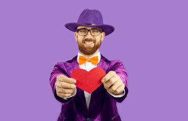 Positive man wearing purple glamorous suit, hat and glasses holding paper red heart and looking at camera with playful eyes, holding symbol of love. Indoor studio shot isolated on purple background.