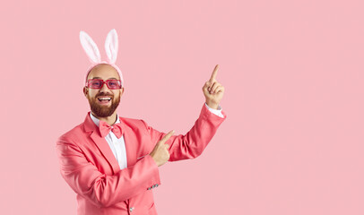 Happy handsome man with ginger beard, in suit, party glasses and cute funny Easter Bunny ears standing on pink background points to side asking to look at something here on blank advertising copyspace