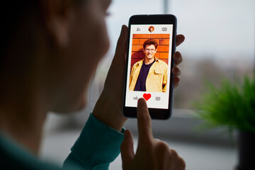 Portrait of a smiling young woman putting red heart button giving like to a cheerful handsome man on her mobile dating application. Find love and online dating concept. Selective focus. - obrazy, fototapety, plakaty