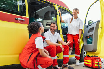 Multi-ethnic group of three paramedics at the rear of an ambulance, climbing in through the open doors. The two women are smiling at the camera, and their male colleague has a serious expression.