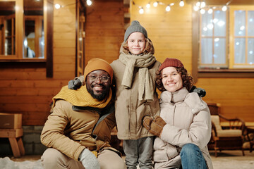 Fototapeta na wymiar Cheerful intercultural mother and father in warm winterwear surrounding their happy cute little son while enjoying winter vacation