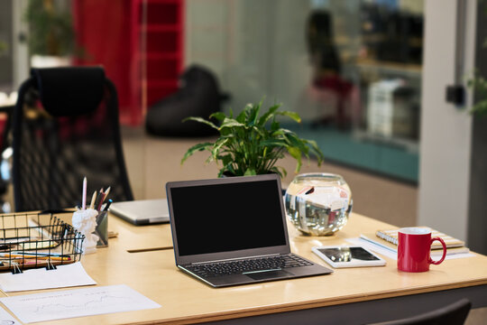 Laptop with black screen standing on desk with green plant in flowerpot, red mug, tablet, spherical fish tank with water and other stuff - Powered by Adobe