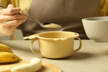 Healthy baby food. Woman putting delicious banana puree into bowl at light wooden table, closeup