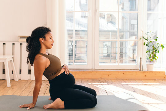Side view of african american pregnant female sitting on floor with legs bend, leaning backwards on hand, relaxing back, meditating with closed eyes, touching belly feeling pushes of her baby