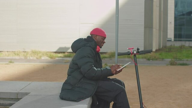 young african american man in streetwear using tablet sitting on electric scooter