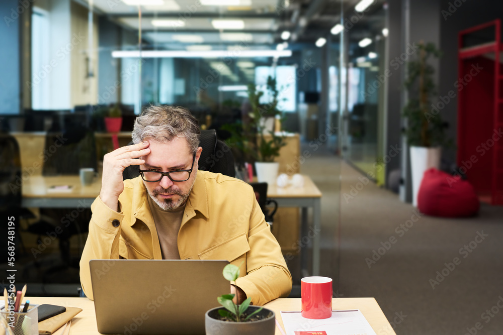 Poster serious mature office worker touching head while sitting by desk in front of laptop and looking thro