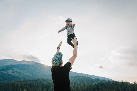 Father Throws Up His Happy Son Into The Sky With His Hands. Walking In Nature On An Autumn Day. Dad And Baby Child Playing In The Mountains. Concept Of Family Spending Time Together On Vacation.