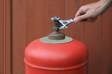 Man with adjustable wrench opening red gas cylinder near brown wooden wall, closeup