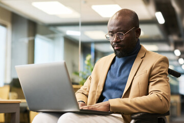 Bearded African American employee with laptop on his knees sitting in wheelchair and looking at screen while networking in office