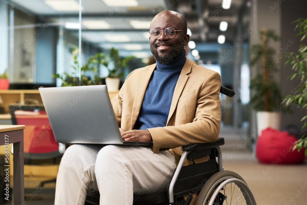 Poster happy young black man with disability sitting in wheelchair in office and looking at camera while an